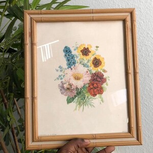 May include: A rectangular picture frame with a light brown bamboo-style border containing a botanical print. The print features a central bouquet of colorful flowers, including yellow, pink, blue, and deep red blossoms with green leaves on a cream background.