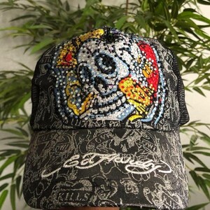 May include: A black trucker hat featuring a prominent skull graphic embellished with colorful rhinestones on the front panel. The fabric displays a repeating grey pattern and the Ed Hardy logo embroidered in white on the brim with the word KILLS.