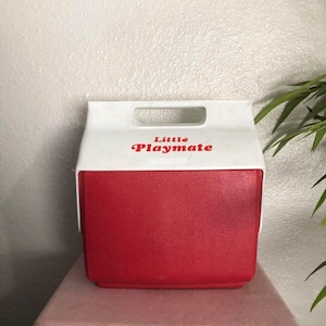 May include: A red and white Little Playmate cooler with a handle. The cooler has a red body and a white top with the words "Little Playmate" in red lettering. The cooler is sitting on a pink surface.