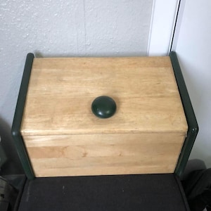 May include: A wooden bread bin with a light wood finish and a dark green knob. The bin has dark green trim and is on a black surface. The rectangular bin has a simple design and is made of wood.