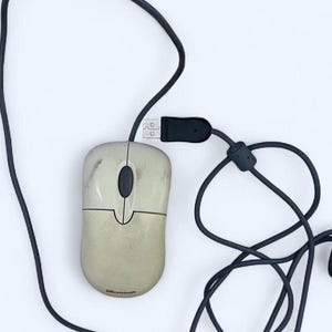 May include: A beige Microsoft computer mouse with a dark gray scroll wheel and a long black USB cable. The mouse features a two-button design with a visible Microsoft logo at the bottom and shows signs of discoloration on the plastic casing.