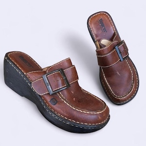 Pode incluir: Um par de tamancos de couro castanho com salto cunha. Os tamancos têm fecho de fivela e costura branca. A marca "Born" é visível na palmilha. Os sapatos estão sobre um fundo branco.