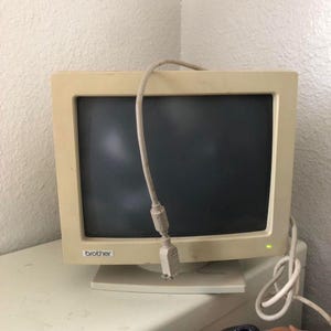 May include: A beige Brother computer monitor with a dark screen and a green power indicator light. A light-colored cable with a connector hangs over the top of the monitor frame, and a coiled cable is visible on the right side of the base.