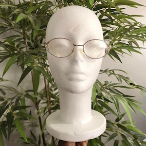 May include: Oval-shaped eyeglasses with a thin, gold-colored metal frame. The glasses are displayed on a white mannequin head, with green leafy plants in the background. The lenses are clear.