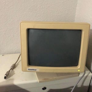 May include: A vintage GoldStar computer monitor with a beige frame and a dark screen. The monitor has a rectangular shape and a small, white GoldStar logo on the bottom left. A cable with a connector extends from the back.