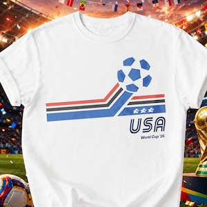 May include: A white short-sleeved t-shirt featuring a graphic of a blue soccer ball with red, white, and blue horizontal stripes. The design includes the text USA and World Cup '26 in blue, along with three small white stars above the text.
