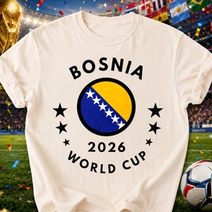May include: A cream-colored t-shirt featuring a circular graphic with the flag of Bosnia and Herzegovina, surrounded by the text "BOSNIA 2026 WORLD CUP" and six black stars arranged in a semi-circle on each side of the central circular emblem.