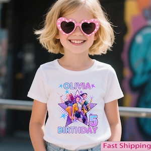 Puede incluir: Una ni&ntilde;a peque&ntilde;a con gafas de sol rosas en forma de coraz&oacute;n y una camiseta blanca. La camiseta presenta un dise&ntilde;o gr&aacute;fico con el nombre "Olivia", el n&uacute;mero "5" y la palabra "Cumplea&ntilde;os". El dise&ntilde;o incluye una estrella e im&aacute;genes de tres chicas.