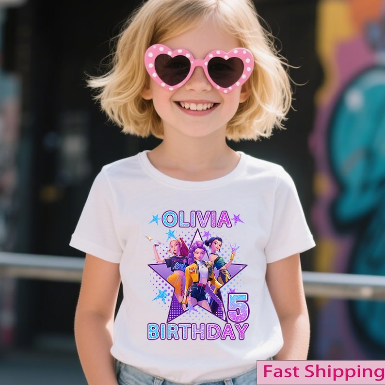 Puede incluir: Una ni&ntilde;a peque&ntilde;a con gafas de sol rosas en forma de coraz&oacute;n y una camiseta blanca. La camiseta presenta un dise&ntilde;o gr&aacute;fico con el nombre "Olivia", el n&uacute;mero "5" y la palabra "Cumplea&ntilde;os". El dise&ntilde;o incluye una estrella e im&aacute;genes de tres chicas.