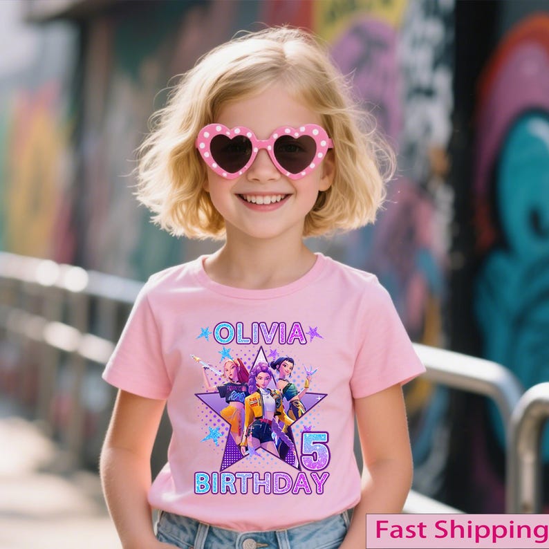 Puede incluir: Una ni&ntilde;a peque&ntilde;a con gafas de sol rosas en forma de coraz&oacute;n y una camiseta rosa. La camiseta presenta el nombre "Olivia", un gr&aacute;fico de personajes animados, el n&uacute;mero "5" y la palabra "Cumplea&ntilde;os". El fondo es una pared de graffiti colorida.