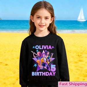 Puede incluir: Una sudadera negra con un gr&aacute;fico colorido de tres personajes animados dentro de una estrella p&uacute;rpura. El texto "OLIVIA" y "BIRTHDAY 5" se muestran. Una ni&ntilde;a peque&ntilde;a lleva la sudadera en una playa.