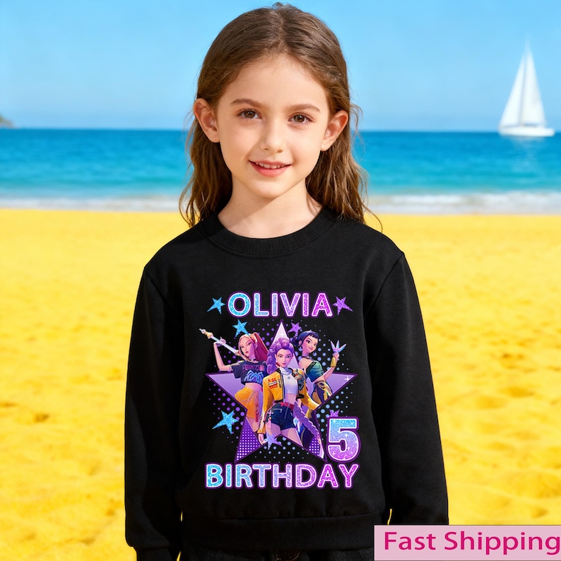 Puede incluir: Una sudadera negra con un gr&aacute;fico colorido de tres personajes animados dentro de una estrella p&uacute;rpura. El texto "OLIVIA" y "BIRTHDAY 5" se muestran. Una ni&ntilde;a peque&ntilde;a lleva la sudadera en una playa.