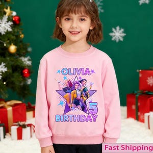 Puede incluir: Sudadera rosa con el nombre "OLIVIA" y el n&uacute;mero "5" impresos. La sudadera presenta un gr&aacute;fico de tres personajes animados dentro de una estrella p&uacute;rpura, con la palabra "BIRTHDAY" debajo.