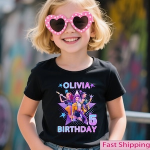 Puede incluir: Una camiseta negra con el nombre "Olivia" y la palabra "Cumplea&ntilde;os" en un dise&ntilde;o colorido. El gr&aacute;fico incluye tres personajes de dibujos animados y el n&uacute;mero "5". La ni&ntilde;a lleva gafas de sol rosas en forma de coraz&oacute;n.