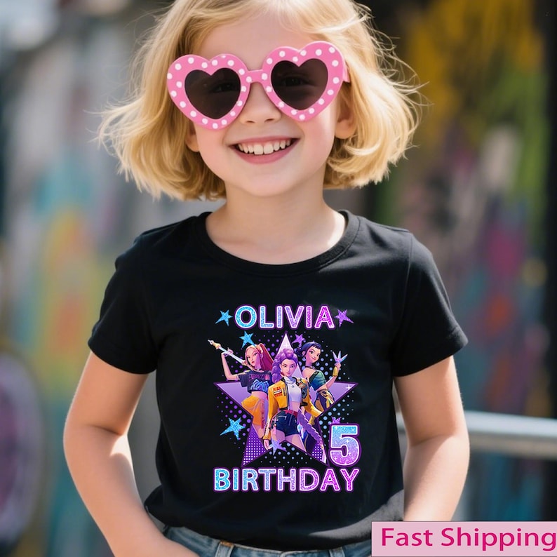 Puede incluir: Una camiseta negra con el nombre "Olivia" y la palabra "Cumplea&ntilde;os" en un dise&ntilde;o colorido. El gr&aacute;fico incluye tres personajes de dibujos animados y el n&uacute;mero "5". La ni&ntilde;a lleva gafas de sol rosas en forma de coraz&oacute;n.
