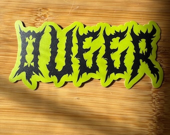 May include: A sticker with the word "HUEGER" in black, stylized, jagged lettering. The text is set against a bright neon green background. The sticker is rectangular and placed on a wooden surface.