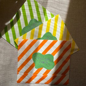 Può includere: Tre sacchetti di carta colorati con strisce diagonali verdi, gialle e arancioni. Ogni sacchetto ha un'etichetta verde con le parole "mystery pack" in un carattere fantasioso. I sacchetti sono piegati e sembrano essere sigillati.