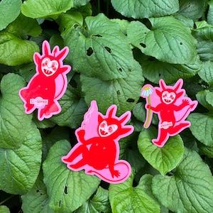 May include: Three red devil stickers with pink accents and cartoonish features. One devil holds a cigarette, one holds a white daisy, and one has a censored bar across its midsection. Each figure has horns, a tail, and a mischievous expression.