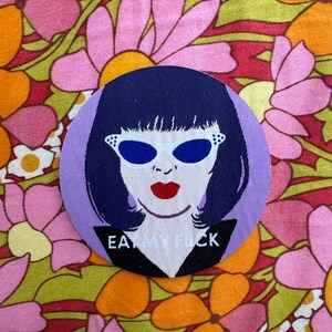 May include: A purple and white embroidered patch featuring a cartoon image of a woman wearing blue sunglasses and red lipstick. The text "EAT MY F*CK" is embroidered in white on a black background.
