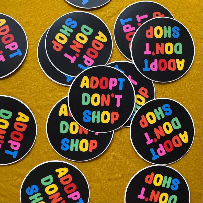 Op de afbeelding: Een verzameling zwarte ronde stickers met de tekst "ADOPT DON'T SHOP" in regenboogkleurige letters. De stickers zijn verspreid over een goudgele ondergrond.