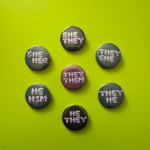 Puede incluir: Siete chapas redondas dispuestas en círculo. Seis chapas son de color gris oscuro con texto iridiscente que dice SHE/THEY, SHE/HER, THEY/SHE, THEY/THEM, THEY/HE, HE/THEY y HE/HIM. La chapa central es de color morado apagado con texto iridiscente: THEY/THEM.