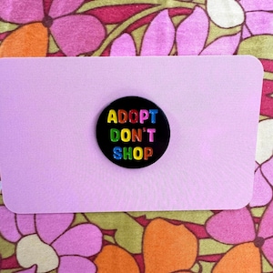 May include: Black button with the text "ADOPT DON'T SHOP" in rainbow colors. The button is on a pink background.