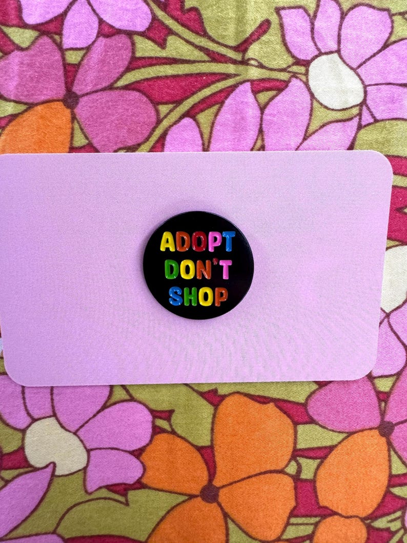 May include: Black button with the text "ADOPT DON'T SHOP" in rainbow colors. The button is on a pink background.