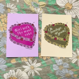 May include: Two greeting cards with heart-shaped cake designs and text. One card is light purple with the text "Girl with the most cake." The other is beige with the text "Oh bondage, up yours!" Both cards have cherry decorations.