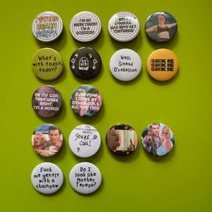 May include: A selection of circular pin badges with different slogans and pictures, arranged on a green surface. The badges display text such as "Psycho Beach Party!" and "Boys spanked, bad boys get tortured!" Some badges have images of people.