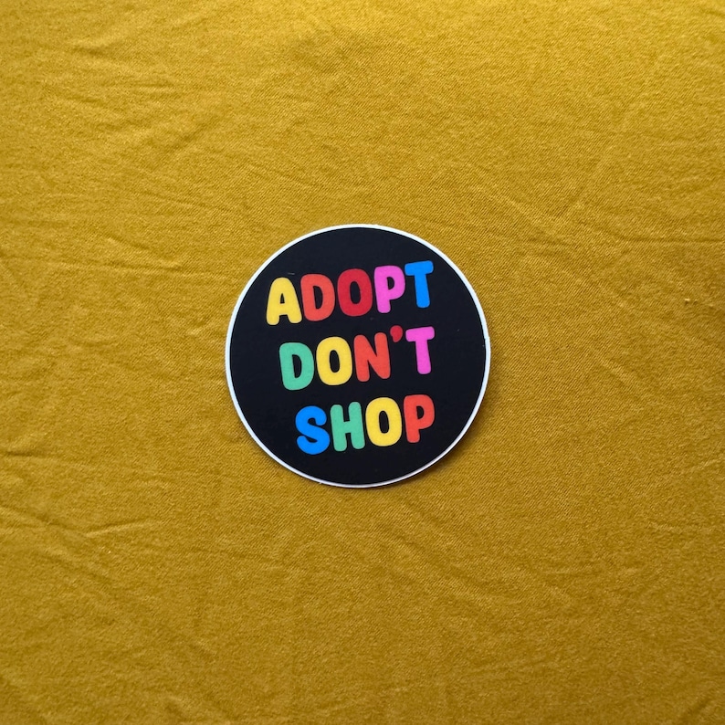 Op de afbeelding: Een ronde sticker met de tekst "ADOPT DON'T SHOP" in regenboogkleuren op een zwarte achtergrond. De sticker is geplaatst op een mosterdgele stof.