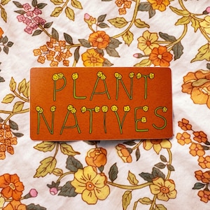 Könnte beinhalten: Ein orangefarbener rechteckiger Aufkleber mit den Worten "PLANT NATIVES" in grünen, floral verzierten Buchstaben. Der Aufkleber befindet sich auf einem Stoff mit floralem Muster mit orangefarbenen, gelben und rosa Blumen.
