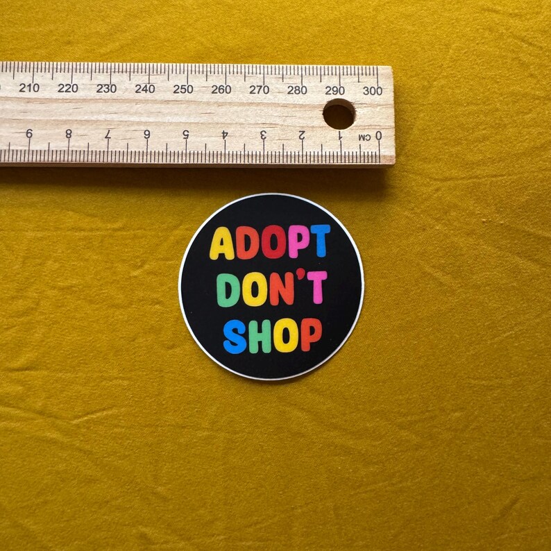 Op de afbeelding: Een ronde zwarte sticker met de tekst "ADOPT DON'T SHOP" in kleurrijke letters. Een houten liniaal met centimeter markeringen ligt boven de sticker op een mosterdgele ondergrond.