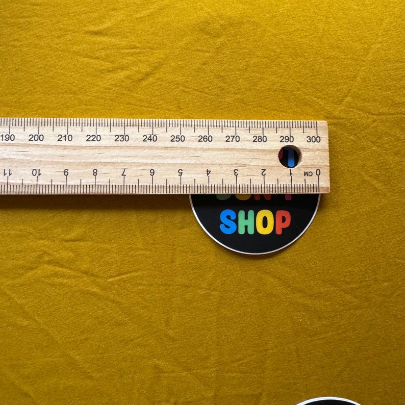 Op de afbeelding: Een houten liniaal met centimeter markeringen, vari&euml;rend van 0 tot 11 cm en van 190 tot 300 cm. De liniaal ligt op een mosterdkleurig oppervlak. Een zwart-witte sticker met het woord "SHOP" staat onderaan.