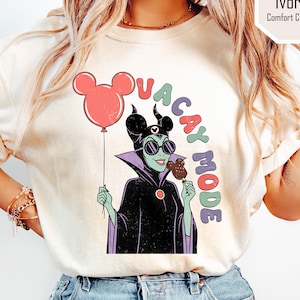 K&ouml;nnte beinhalten: Cremefarbenes T-Shirt mit einer Cartoon-Illustration einer Figur, die einen Mickey-Mouse-Ballon und einen Schokoladen-Snack h&auml;lt. Der Text "VACAY MODE" steht &uuml;ber der Figur. Das Shirt hat einen Rundhalsausschnitt und kurze &Auml;rmel.