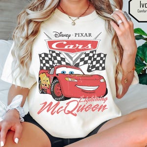 Puede incluir: Camiseta de color marfil con un dise&ntilde;o de Rayo McQueen de Cars de Disney Pixar. El dise&ntilde;o incluye el logotipo de Cars, banderas a cuadros y el texto "Lightning McQueen". La camiseta est&aacute; etiquetada como "Comfort Colors" y "Tee Designed by Megan".