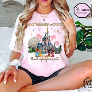 May include: A pale pink t-shirt with a vibrant graphic of Walt Disney World. The design includes Cinderella's castle, the Epcot ball, fireworks, and Disney characters. Text reads "WALT DISNEY WORLD" and "The most magical place on earth."