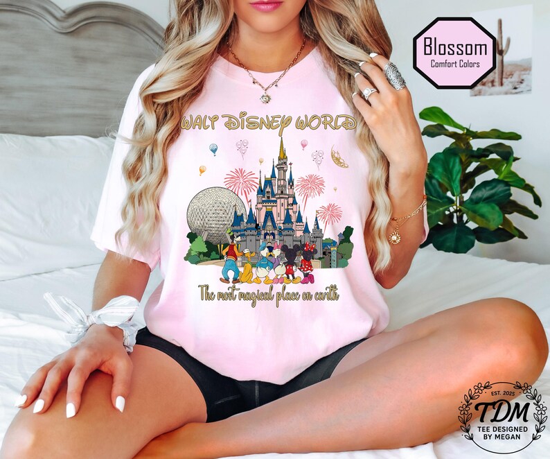 May include: A pale pink t-shirt with a vibrant graphic of Walt Disney World. The design includes Cinderella's castle, the Epcot ball, fireworks, and Disney characters. Text reads "WALT DISNEY WORLD" and "The most magical place on earth."