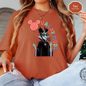 K&ouml;nnte beinhalten: Ziegelrotes T-Shirt mit einer Cartoon-Illustration einer Figur mit Sonnenbrille, die einen Mickey Mouse-Ballon und ein Eis am Stiel h&auml;lt. Die Worte "VACAY MODE" sind vertikal aufgedruckt. Das T-Shirt hat eine lockere Passform.
