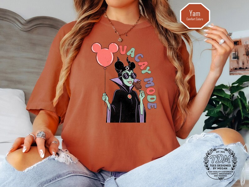 K&ouml;nnte beinhalten: Ziegelrotes T-Shirt mit einer Cartoon-Illustration einer Figur mit Sonnenbrille, die einen Mickey Mouse-Ballon und ein Eis am Stiel h&auml;lt. Die Worte "VACAY MODE" sind vertikal aufgedruckt. Das T-Shirt hat eine lockere Passform.