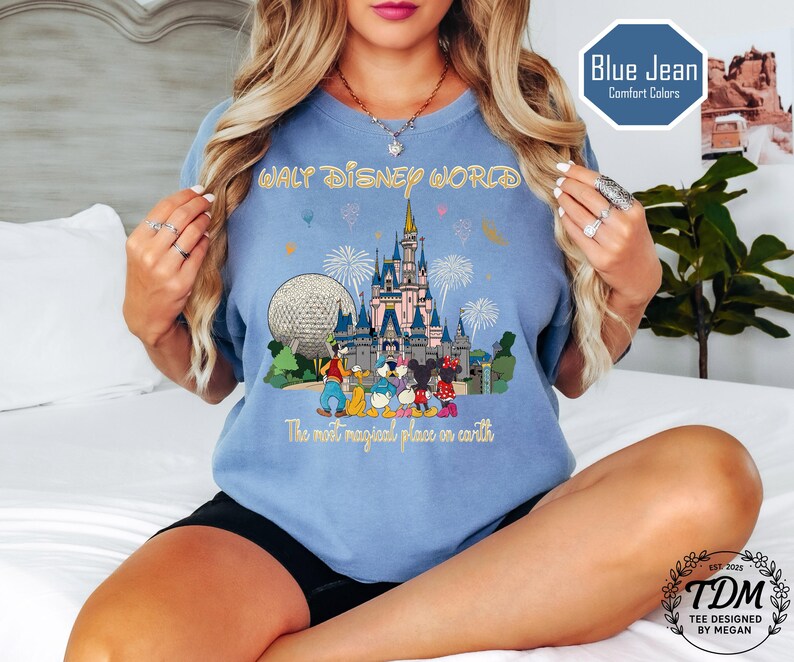 May include: A light blue t-shirt with a colourful graphic of Cinderella's castle, Spaceship Earth, and Disney characters. The text "WALT DISNEY WORLD" and "The most magical place on earth" are printed on the shirt.