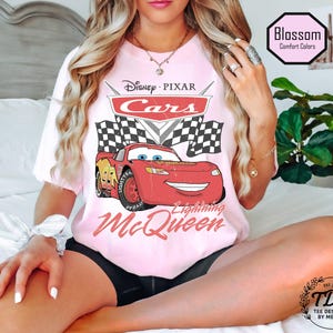 Puede incluir: Una camiseta rosa claro con el logotipo de Disney Pixar Cars y Lightning McQueen. El dise&ntilde;o incluye banderas a cuadros y el texto "Lightning McQueen". La camiseta tambi&eacute;n tiene el texto "Blossom Comfort Colors" y "TDM Tee Designed by Megan".