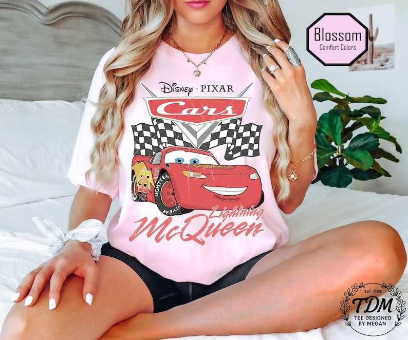 Puede incluir: Una camiseta rosa claro con el logotipo de Disney Pixar Cars y Lightning McQueen. El dise&ntilde;o incluye banderas a cuadros y el texto "Lightning McQueen". La camiseta tambi&eacute;n tiene el texto "Blossom Comfort Colors" y "TDM Tee Designed by Megan".