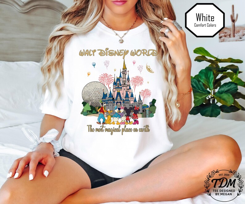 May include: White t-shirt with a colourful graphic of the Walt Disney World castle, characters, and fireworks. The text "WALT DISNEY WORLD" is in gold, with the phrase "The most magical place on earth" below the graphic.