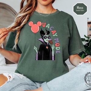 K&ouml;nnte beinhalten: Moosgr&uuml;nes T-Shirt mit einer Cartoon-Illustration eines Disney-B&ouml;sewichts, der einen Mickey-Mouse-Ballon und ein Eis h&auml;lt. Der Text "VACAY MODE" ist in bunten Buchstaben gehalten. Das Shirt ist eine Komfortfarbe.