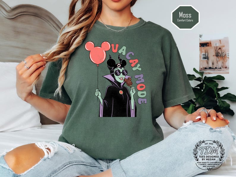 K&ouml;nnte beinhalten: Moosgr&uuml;nes T-Shirt mit einer Cartoon-Illustration eines Disney-B&ouml;sewichts, der einen Mickey-Mouse-Ballon und ein Eis h&auml;lt. Der Text "VACAY MODE" ist in bunten Buchstaben gehalten. Das Shirt ist eine Komfortfarbe.