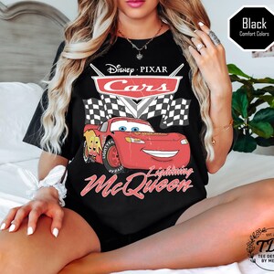 Puede incluir: Camiseta negra con un dise&ntilde;o de Rayo McQueen de Cars de Disney Pixar. El dise&ntilde;o incluye el logotipo de Cars, banderas a cuadros y el texto "Lightning McQueen". La camiseta tambi&eacute;n tiene el texto "TDM Tee Designed by Megan".
