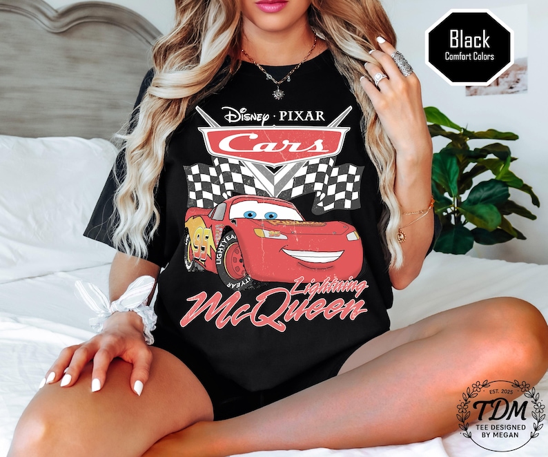 Puede incluir: Camiseta negra con un dise&ntilde;o de Rayo McQueen de Cars de Disney Pixar. El dise&ntilde;o incluye el logotipo de Cars, banderas a cuadros y el texto "Lightning McQueen". La camiseta tambi&eacute;n tiene el texto "TDM Tee Designed by Megan".