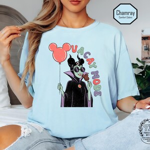 K&ouml;nnte beinhalten: Hellblaues T-Shirt mit einer Cartoon-Illustration einer Figur mit Sonnenbrille, die einen Mickey Mouse-Ballon und ein Eis h&auml;lt. Der Text "VACAY MODE" ist &uuml;ber der Figur angeordnet. Das T-Shirt hat eine lockere Passform.