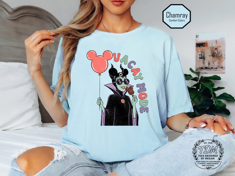 K&ouml;nnte beinhalten: Hellblaues T-Shirt mit einer Cartoon-Illustration einer Figur mit Sonnenbrille, die einen Mickey Mouse-Ballon und ein Eis h&auml;lt. Der Text "VACAY MODE" ist &uuml;ber der Figur angeordnet. Das T-Shirt hat eine lockere Passform.