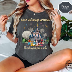 May include: Dark grey t-shirt with a colourful graphic of Cinderella's castle, Spaceship Earth, and fireworks, with the text "WALT DISNEY WORLD" and "The most magical place on earth". The shirt is a Pepper Comfort Colors tee.